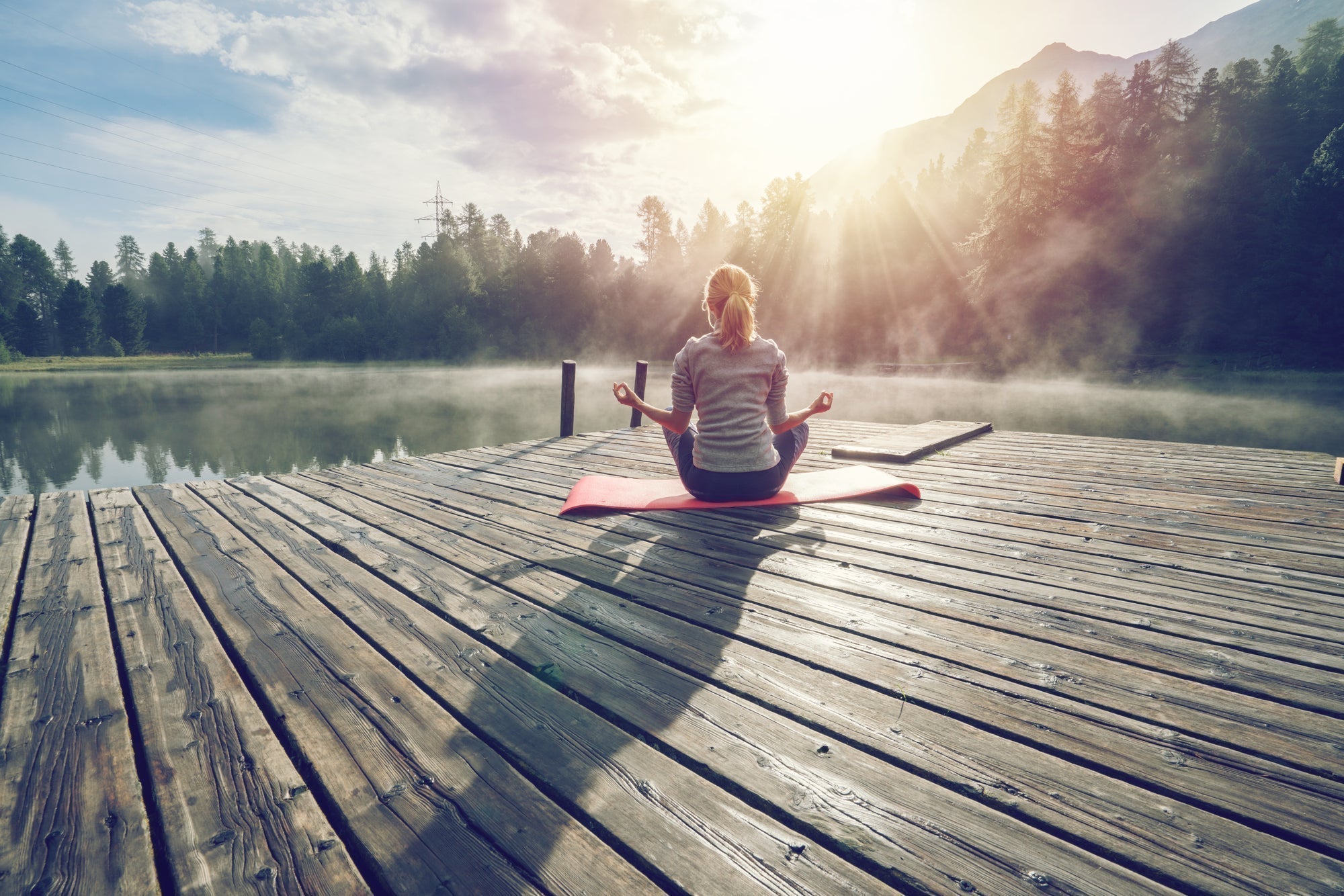 Frau sitzt bei Sonnenaufgang auf einer Yogamatte auf einem Steg am See und meditiert