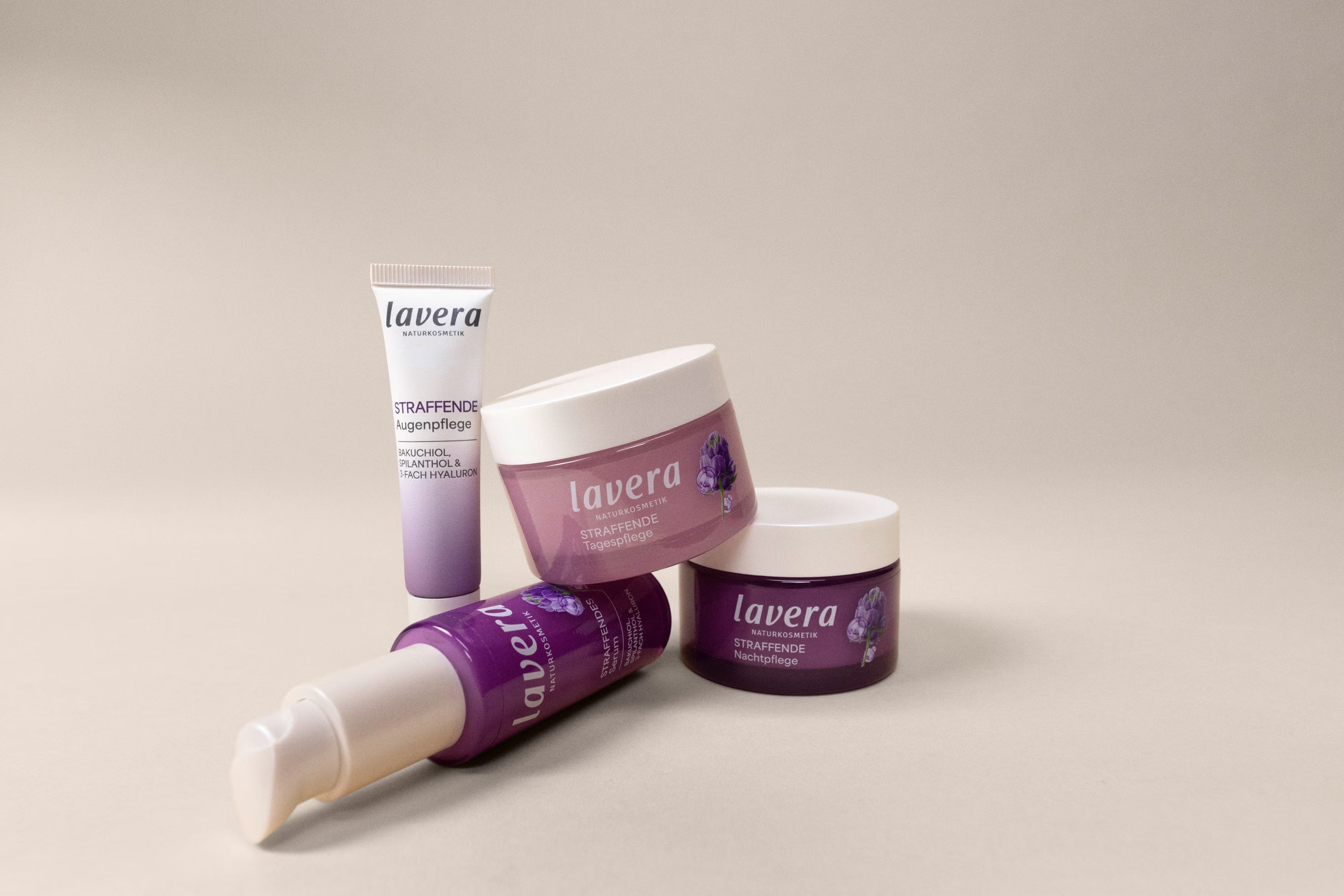 Vier lavera Produkte der straffenden Gesichtspflege in violetten Verpackungen liegen und stehen vor hellem Hintergrund.
