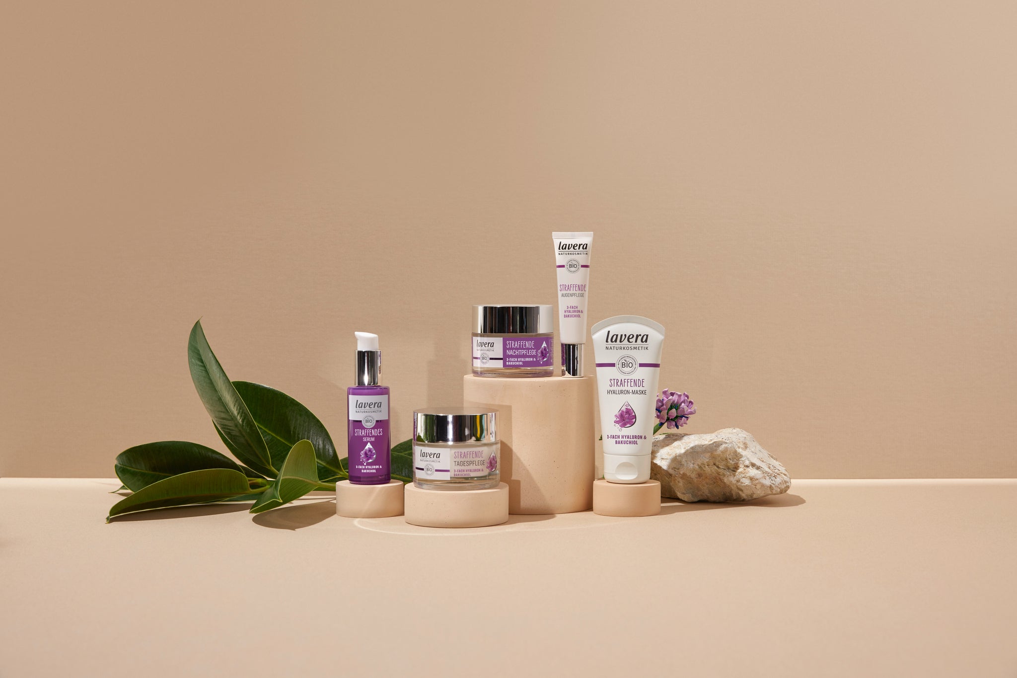 lavera Straffende Anti-Age Pflegeserie mit Hyaluron und Bakuchiol: Serum, Tagescreme, Nachtcreme, Augenpflege und Maske, arrangiert mit Blättern und Stein auf beigem Hintergrund.