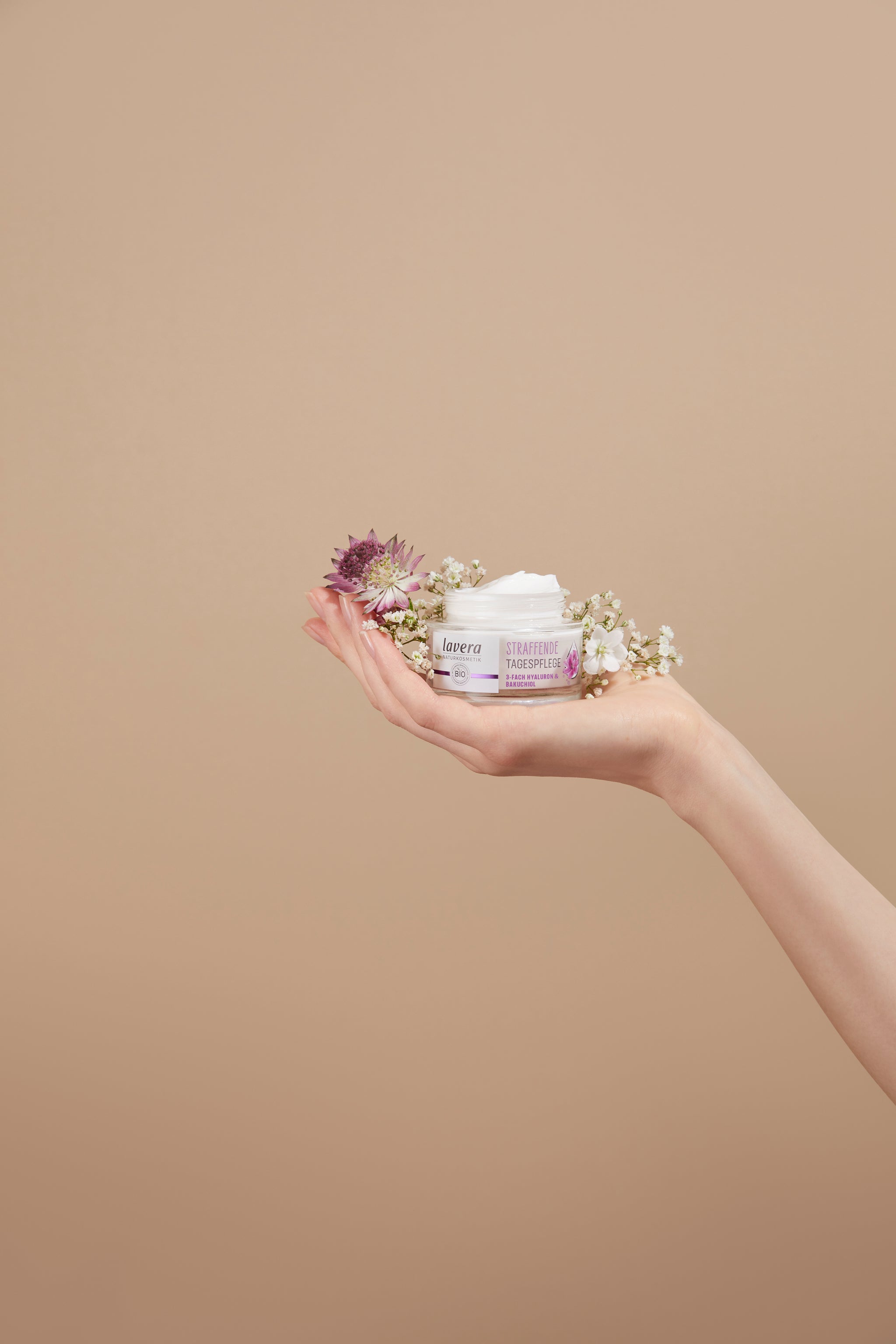 Hand hält Lavera Straffende Tagespflege mit Hyaluron und Bakuchiol, dekoriert mit zarten Blüten vor beigem Hintergrund.