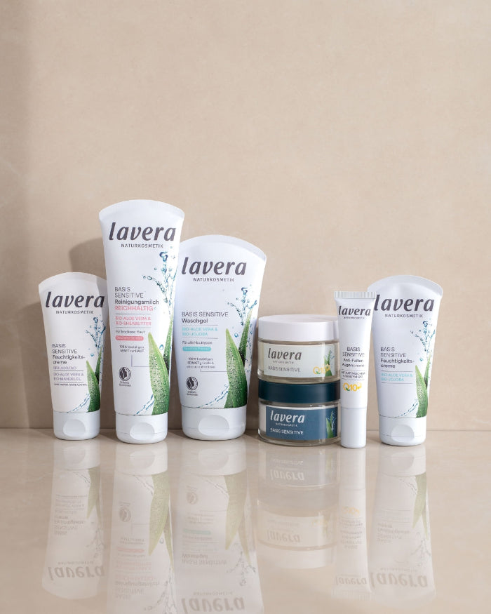 Mehrere lavera BASIS SENSITIVE Pflegeprodukte für das Gesicht stehen nebeneinander auf einer reflektierenden Oberfläche.