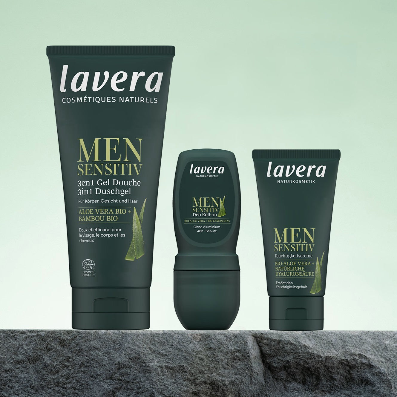 Drei lavera MEN SENSITIVE Pflegeprodukte im Set – 3-in-1-Duschgel, Deo-Roll-on und Feuchtigkeitspflege – stehen vor hellgrünem Hintergrund auf einem Stein.