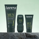 Drei lavera MEN SENSITIVE Pflegeprodukte im Set – 3-in-1-Duschgel, Deo-Roll-on und Feuchtigkeitspflege – stehen vor hellgrünem Hintergrund auf einem Stein.