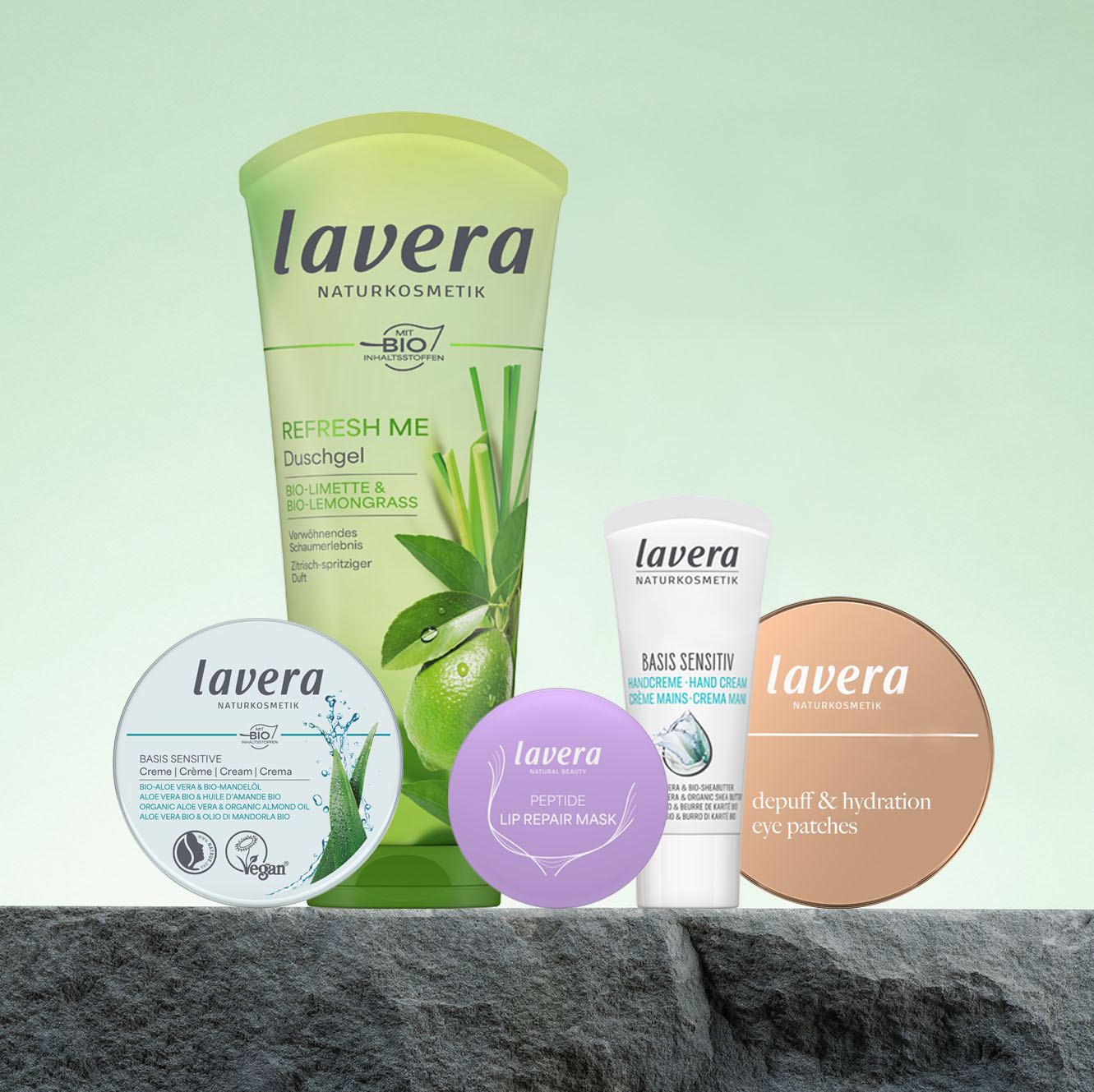 Mehrere lavera Pflegeprodukte im Set, darunter ein Refresh-Me-Duschgel, eine Gesichtscreme, eine Lippenmaske und Augenpads, stehen vor hellgrünem Hintergrund auf einem Stein.
