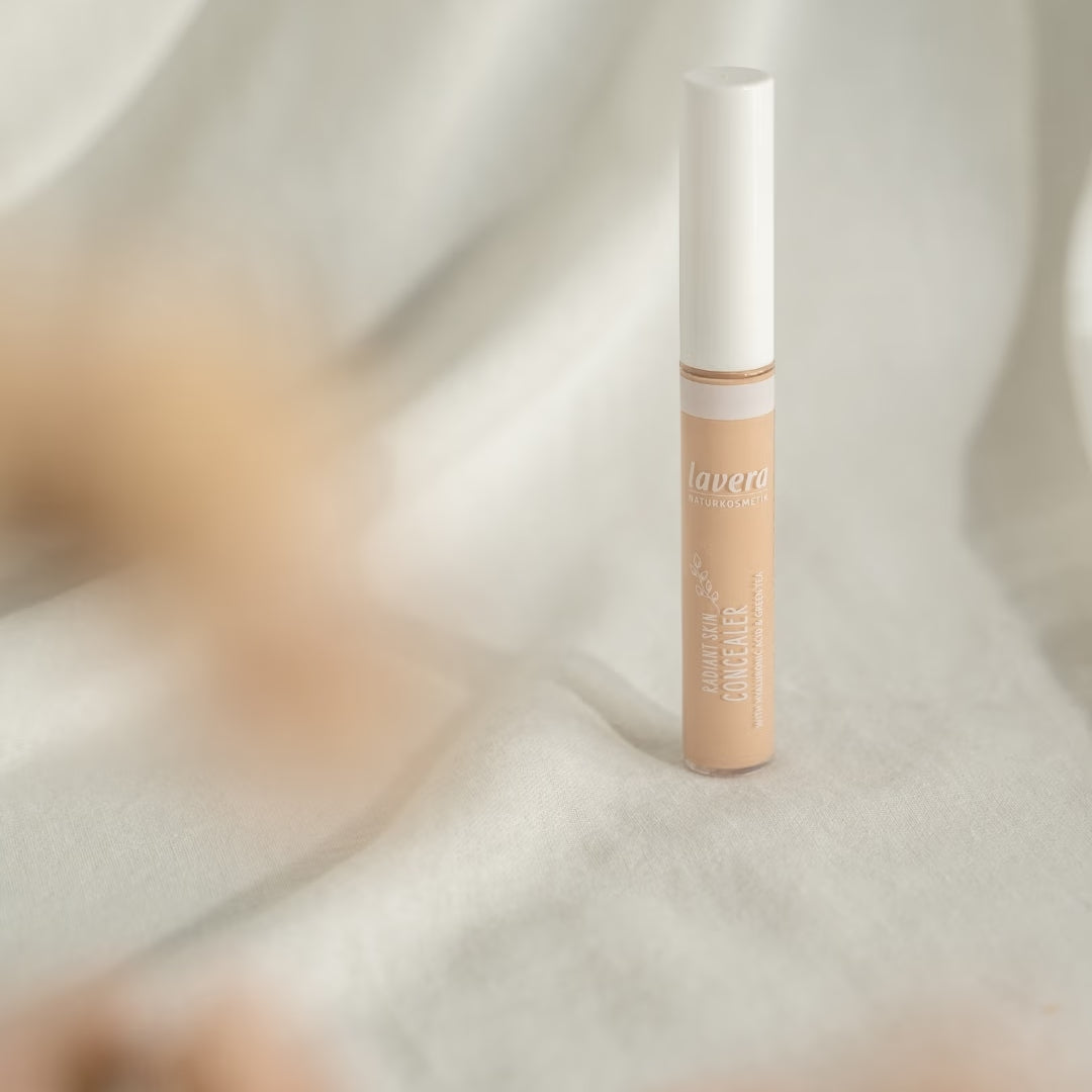 Ein lavera Liquid Skin Concealer steht auf hellem Stoff vor einem weich drapierten Hintergrund mit unscharfen beigefarbenen Akzenten.