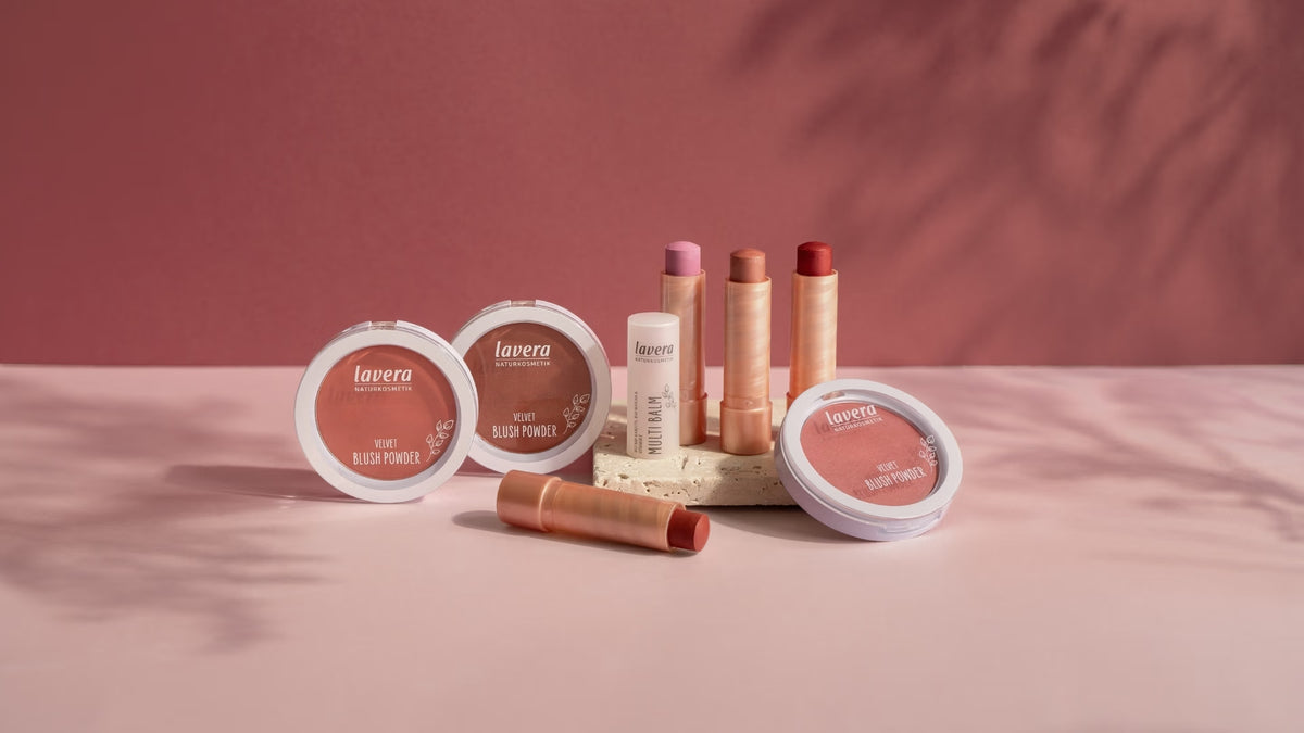 Mehrere lavera Rouge-Produkte und Multi Balms in Rosé-, Nude- und Rottönen stehen auf einem Podest vor rosafarbenem Hintergrund.