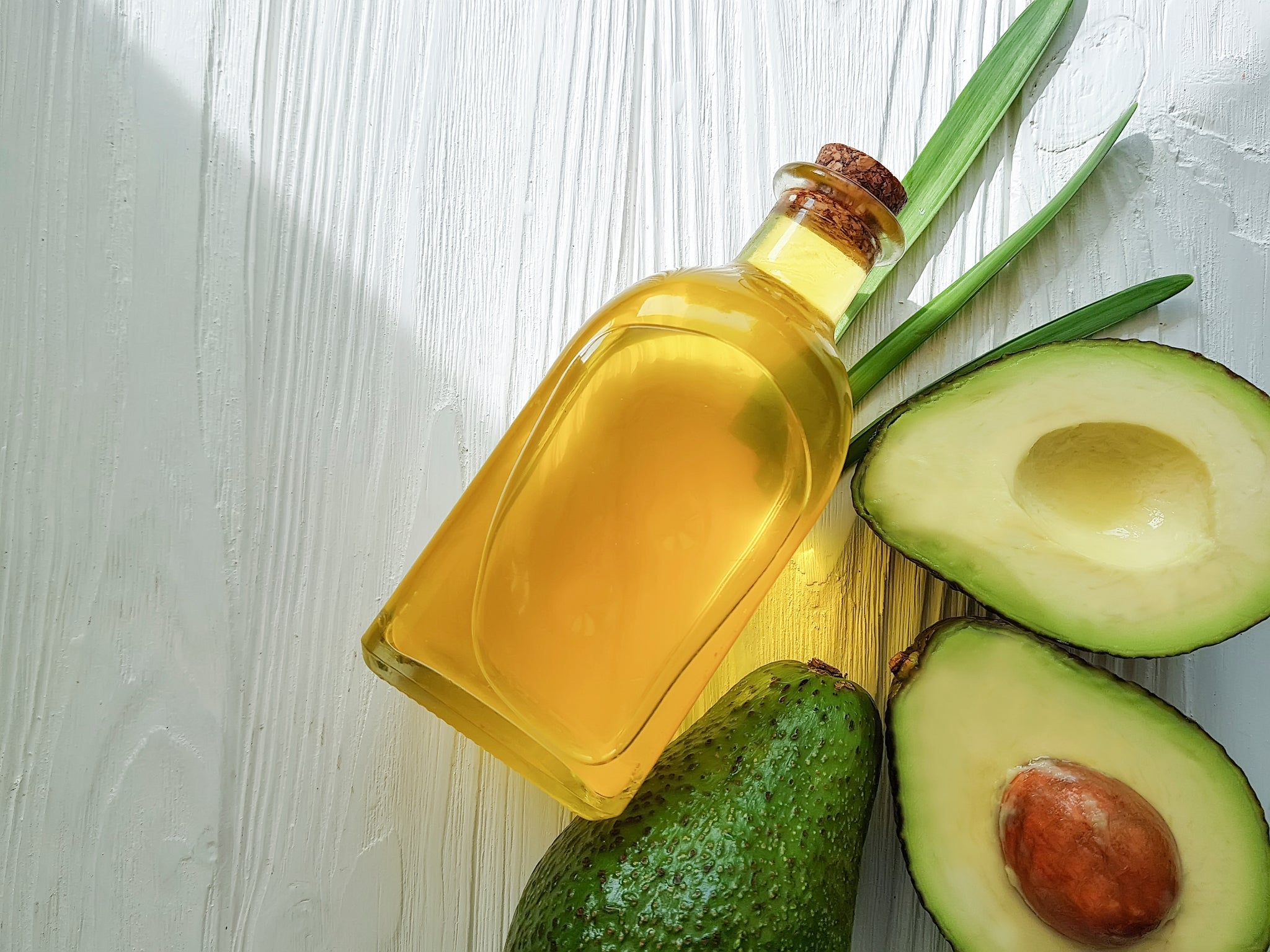 Glasflasche mit goldenem Avocadoöl neben einer ganzen und halbierten Avocado auf hellem Holztisch.