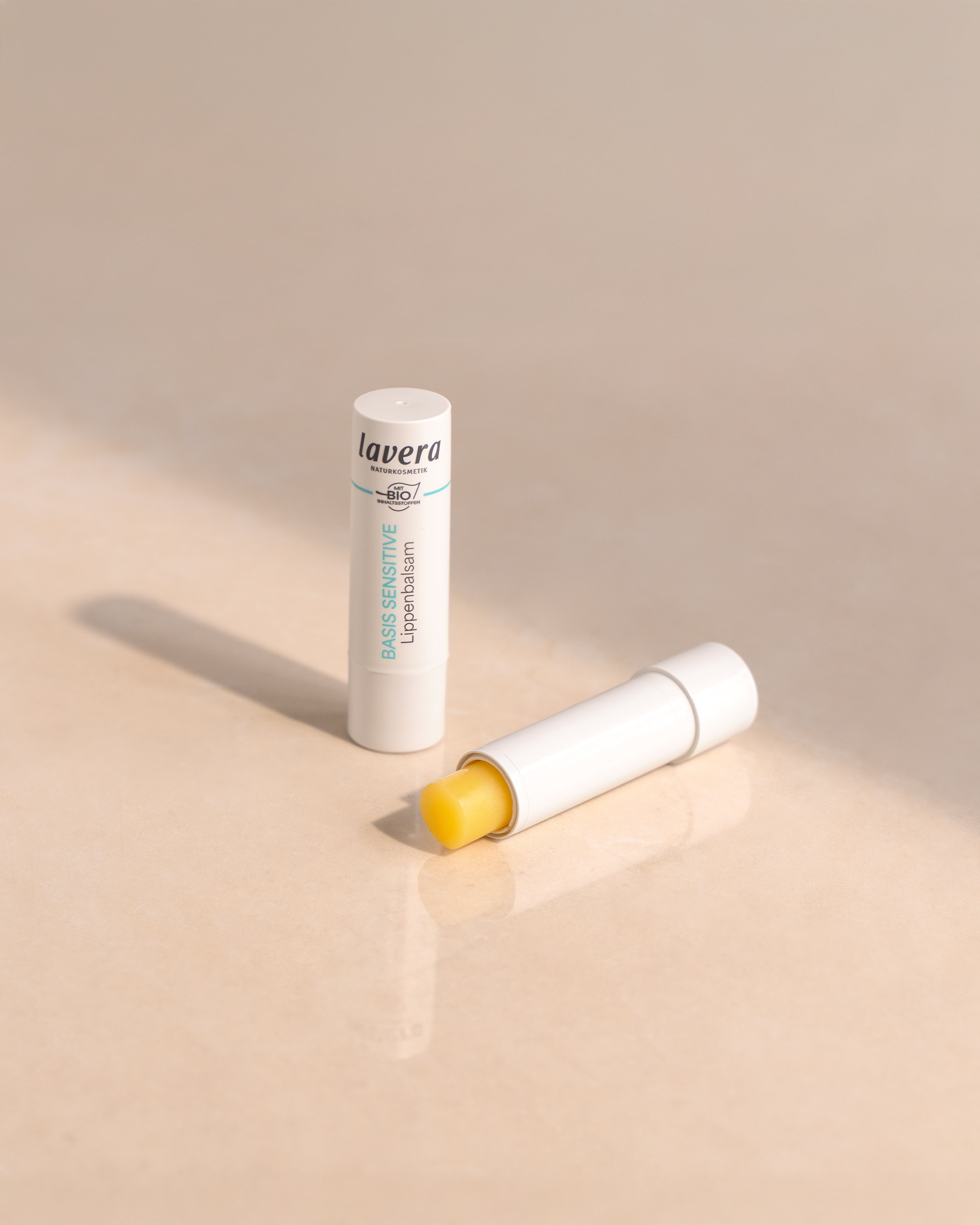 lavera Basis Sensitive Lippenbalsam in weißer Verpackung, geöffnet und geschlossen auf hellem Hintergrund.