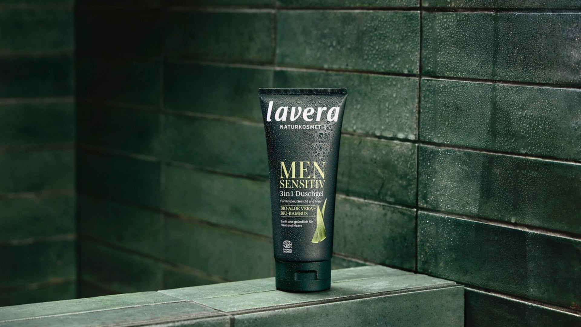 Eine Tube lavera MEN SENSITIVE 3-in-1-Duschgel steht mit Wassertropfen vor dunkelgrünen Fliesen.
