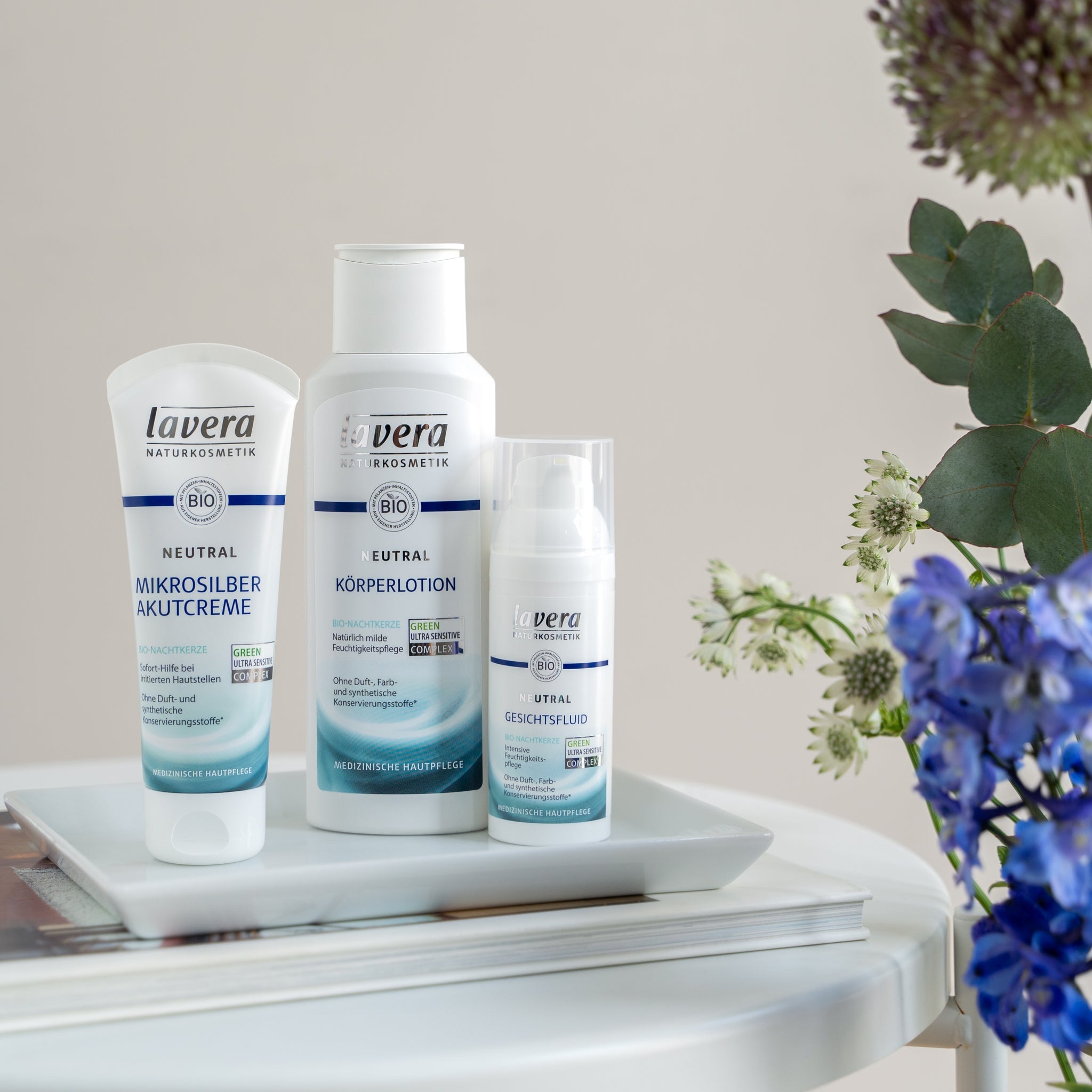Lavera Neutral medizinische Hautpflegeprodukte mit Mikrosilber: Akutcreme, Körperlotion und Gesichtsfluid, dekorativ mit Blumen auf einem weißen Tisch arrangiert.