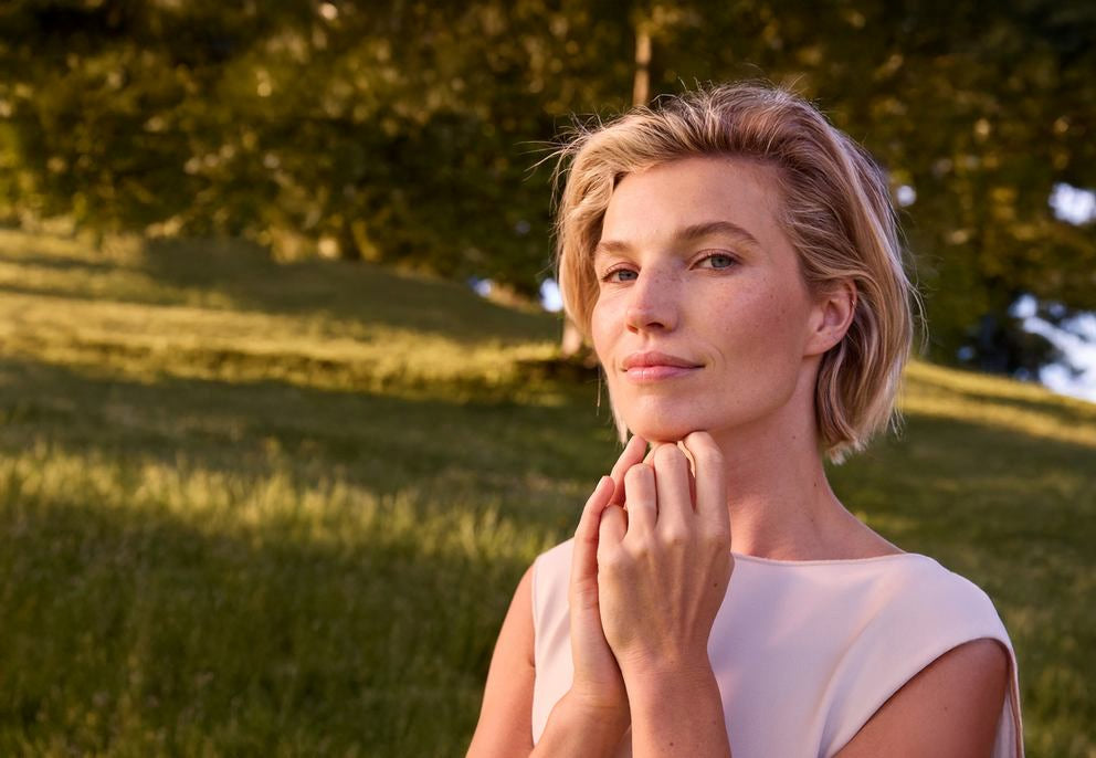 Junge Frau mit blonden Haaren, die vor einer Wiese steht und in die Kamera lächelt