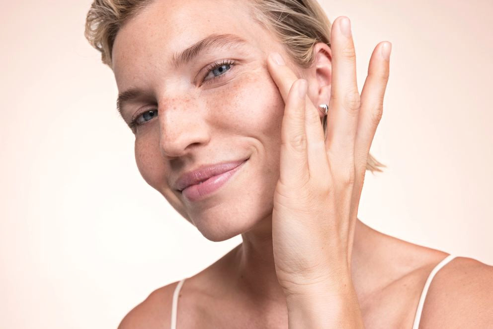 Eine blonde Frau streicht mit einem Finger über ihre Wange und lächelt.