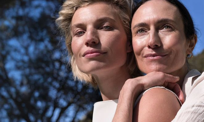 Ein blondes und ein dunkelhaariges Modell stehen vor einem Baum und halten sich aneinander fest.