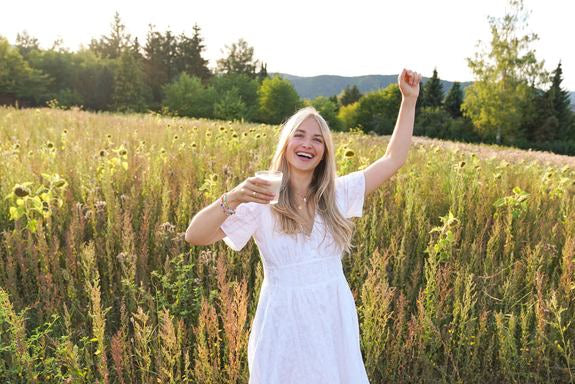 Frau im weißen Kleid lacht auf einer Wiese mit Glas in der Hand