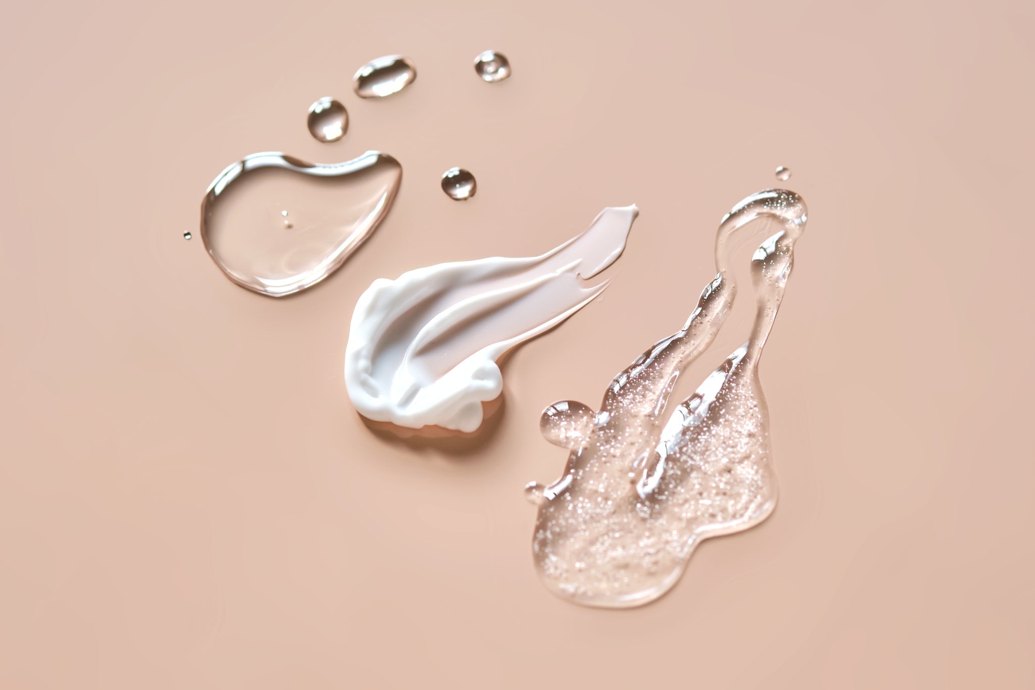Texturen: weiße Creme, glitzerndes Gel und klare Tropfen auf beigem Hintergrund