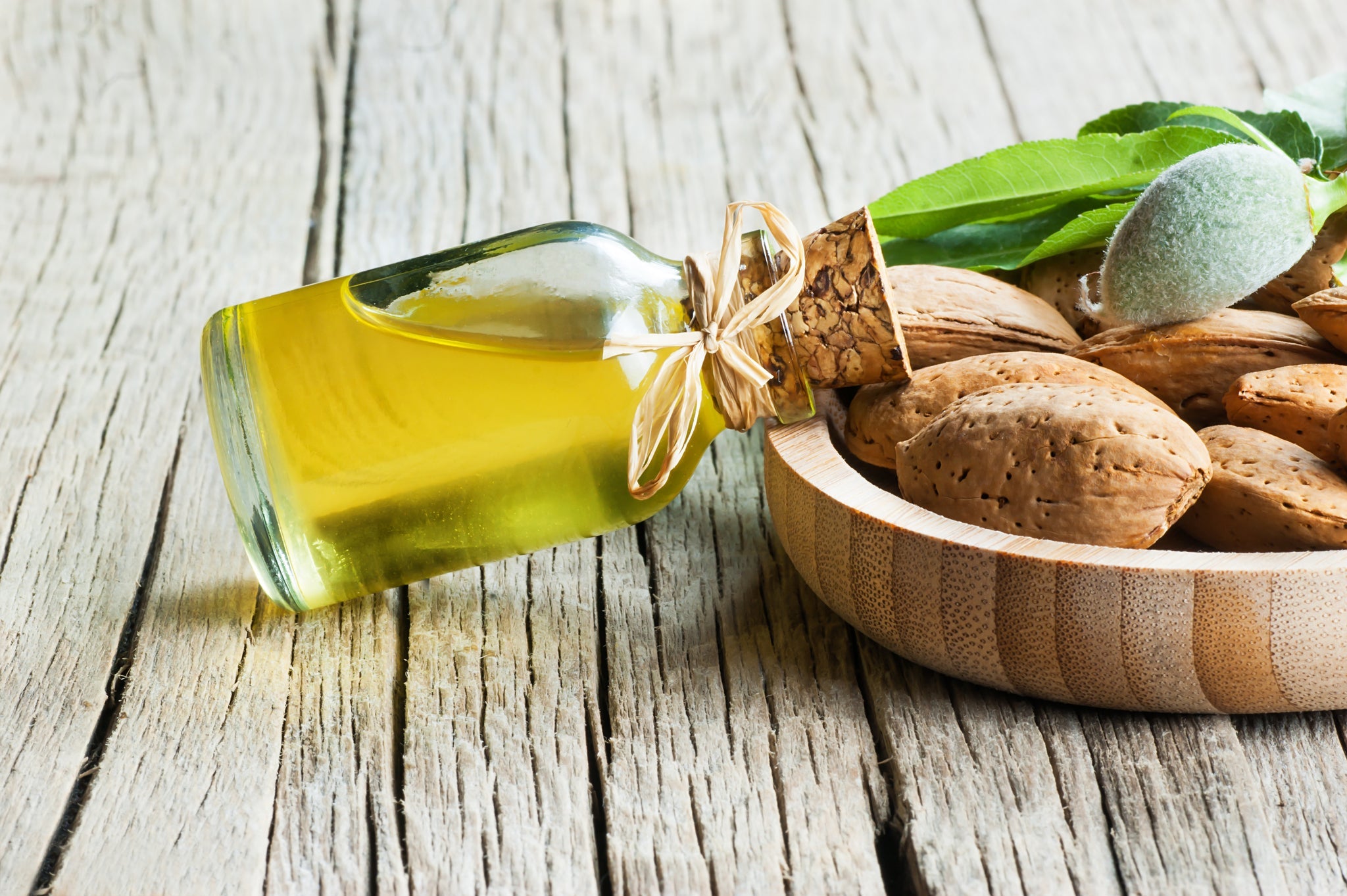 Glasflasche mit goldenem Mandelöl neben einer Holzschale voller Mandeln und grüner Blätter auf Holztisch.
