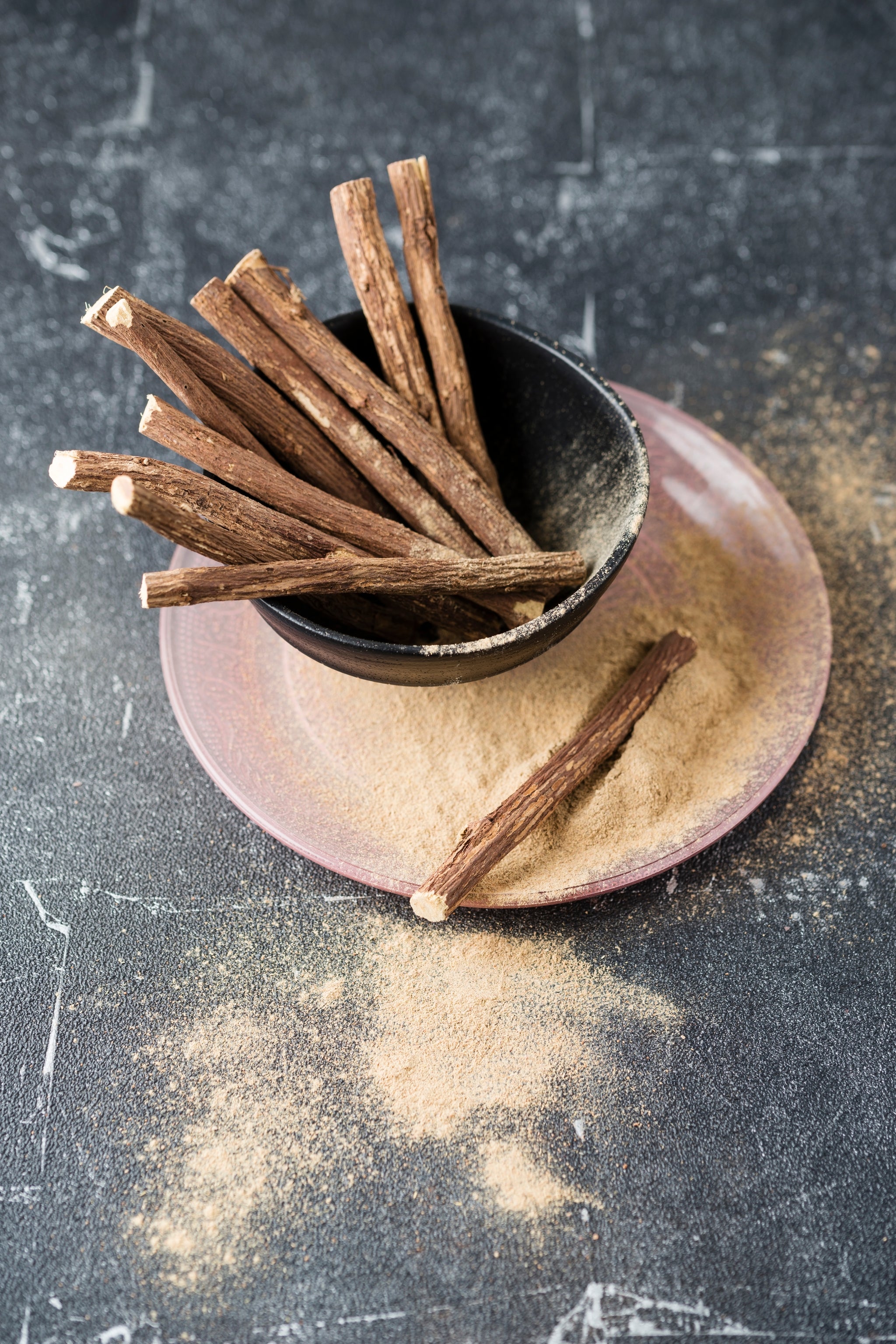 Süßholzwurzeln in einer Schale, daneben Süßholzpulver auf einem Teller verstreut.