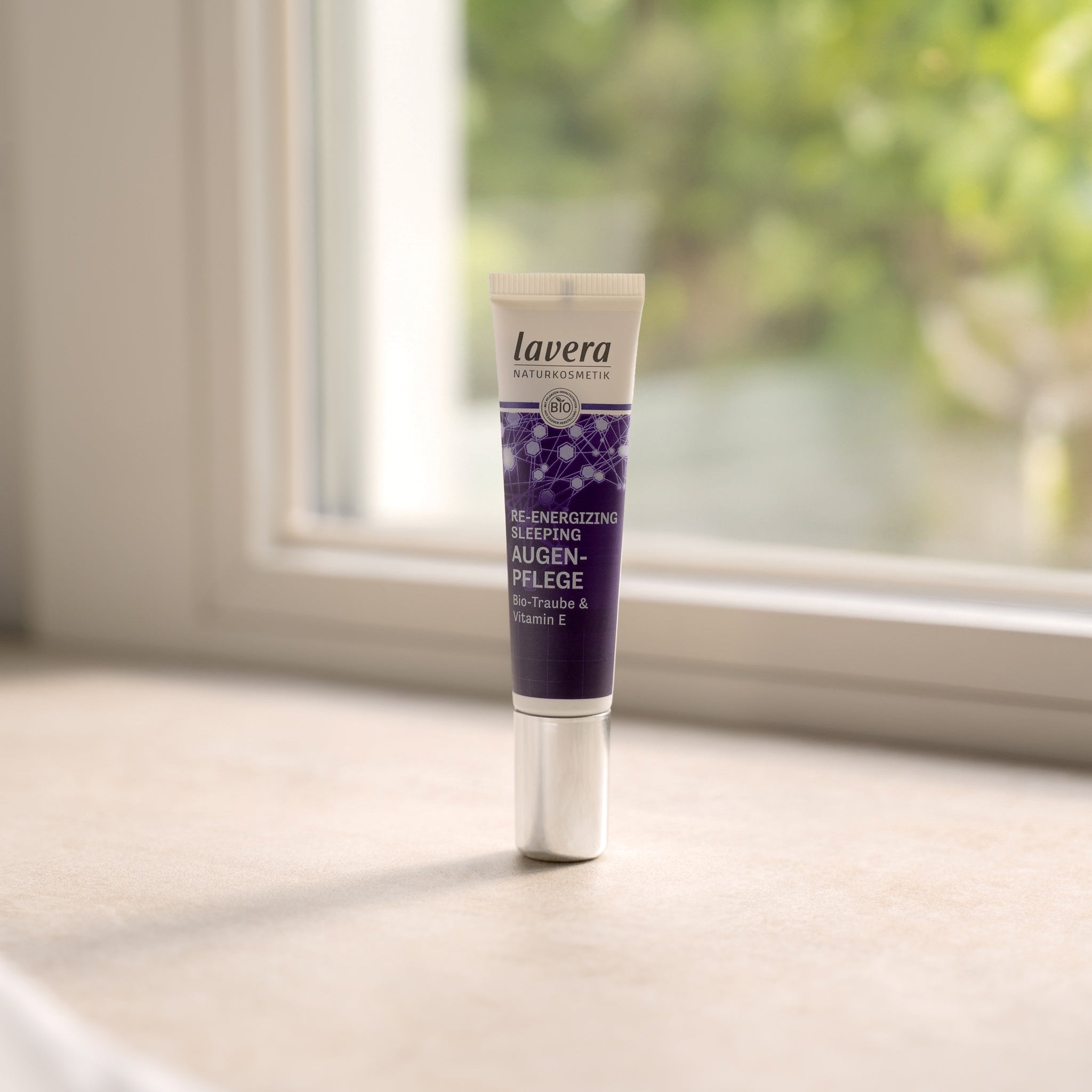 Eine kleine violette Tube mit silbernem Verschluss des lavera Naturkosmetik Re-Energizing Sleeping Augenpflege-Produkts auf einer Fensterbank im Tageslicht platziert, unscharfes Grün im Hintergrund.