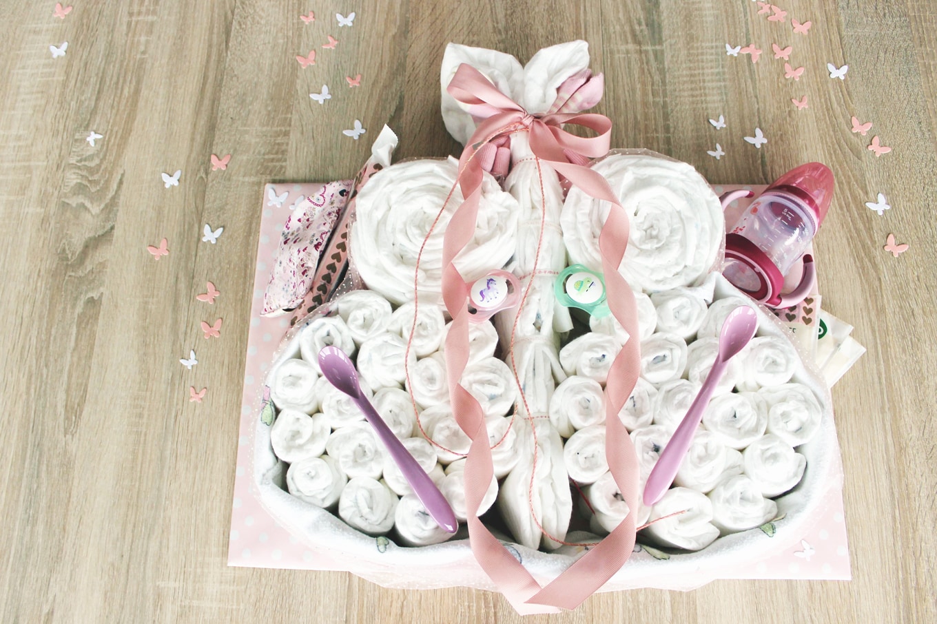 windelschmetterling-geschenk-geburt-diy zusammengerollte weiße Windeln in Form eines schmetterlingsmit baby accessoires und geschenkband auf hellem holztisch